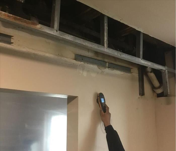 SERVPRO technician in PPE using pinless moisture sensor on a wall near a sealed doorway. The drywall and framing above have b