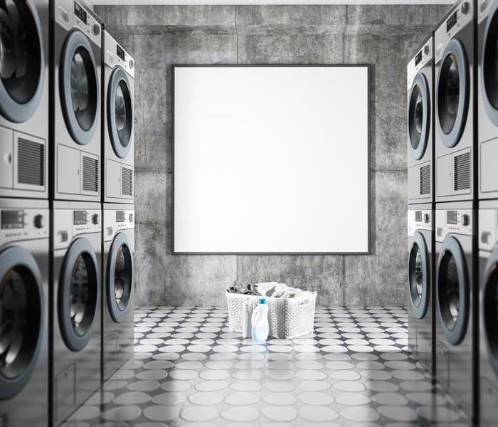 Washing machines and dryers stacked up in a room with a basket of  laundry and detergent on the floor. 